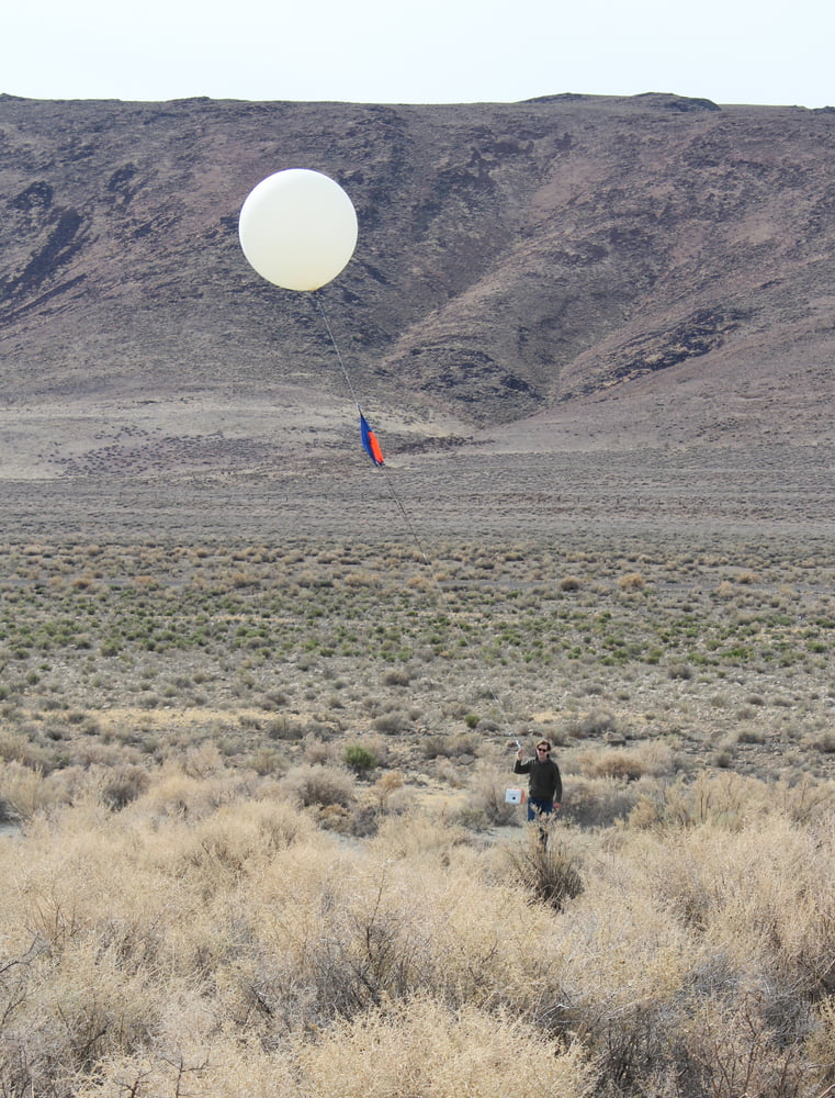 Chris in the distance, in scrubland holding a partially inflated weather balloon attached to a small foam payload